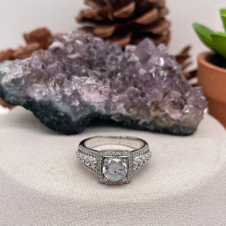 May include: A silver ring with a square-shaped center stone surrounded by smaller stones. The ring is set on a white surface with a purple amethyst crystal behind it.