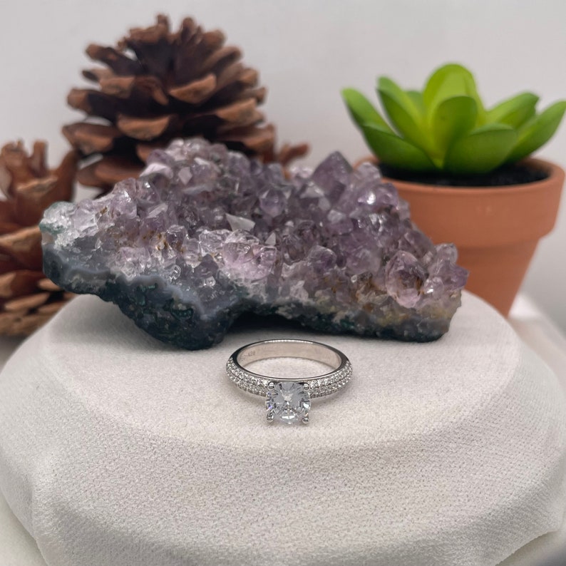 May include: A silver ring with a large, clear, round gemstone surrounded by smaller gemstones. The ring is sitting on a white, textured surface.