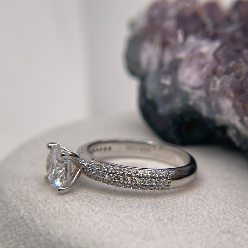 May include: A silver engagement ring featuring a large, clear, round-cut centre stone and a double row of smaller diamonds on the band.