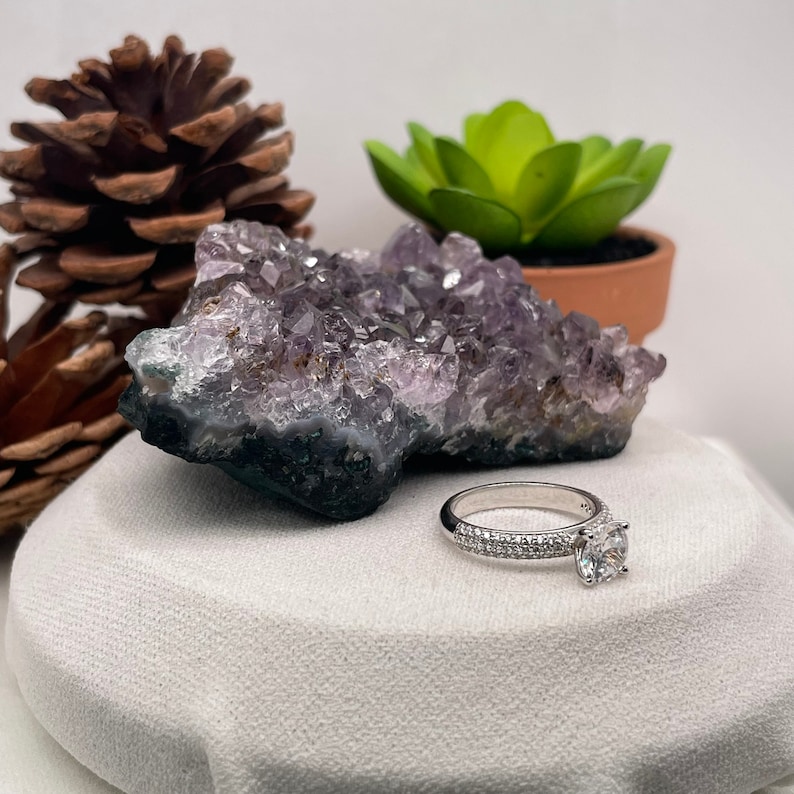 May include: A silver ring with a round, clear gemstone surrounded by smaller gemstones. The ring is on a white surface next to a purple amethyst crystal.