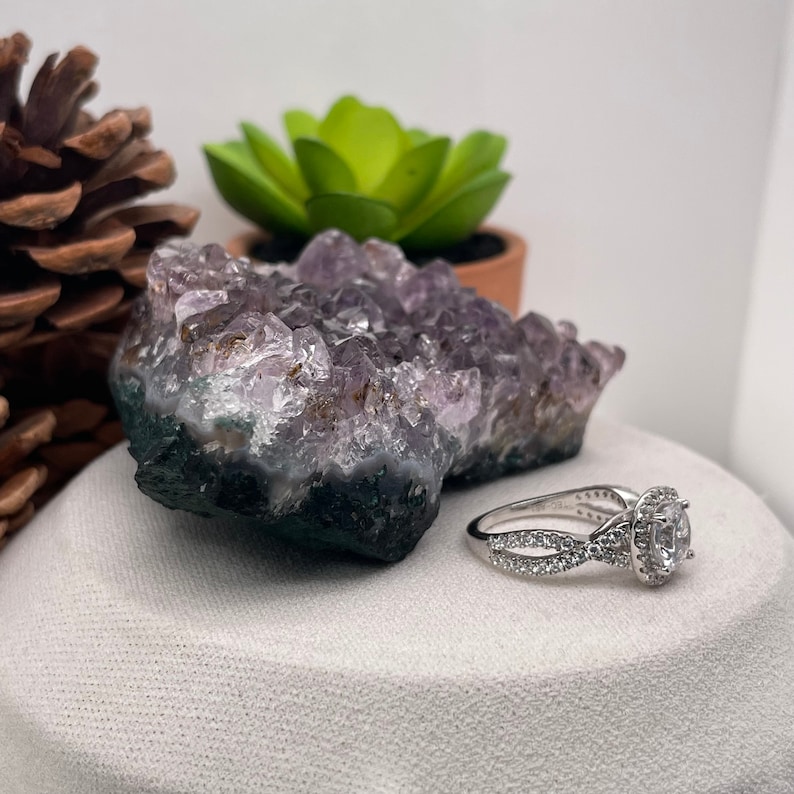 Puede incluir: Un anillo de compromiso de plata con una piedra central redonda y clara rodeada de piedras m&aacute;s peque&ntilde;as. El anillo est&aacute; sobre una superficie blanca junto a un cristal de amatista p&uacute;rpura.