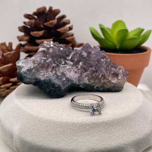 May include: A silver engagement ring featuring a large, round, clear centre stone and smaller stones set in a band. The ring is displayed on a white surface with a purple amethyst crystal and a small green succulent plant in the background.