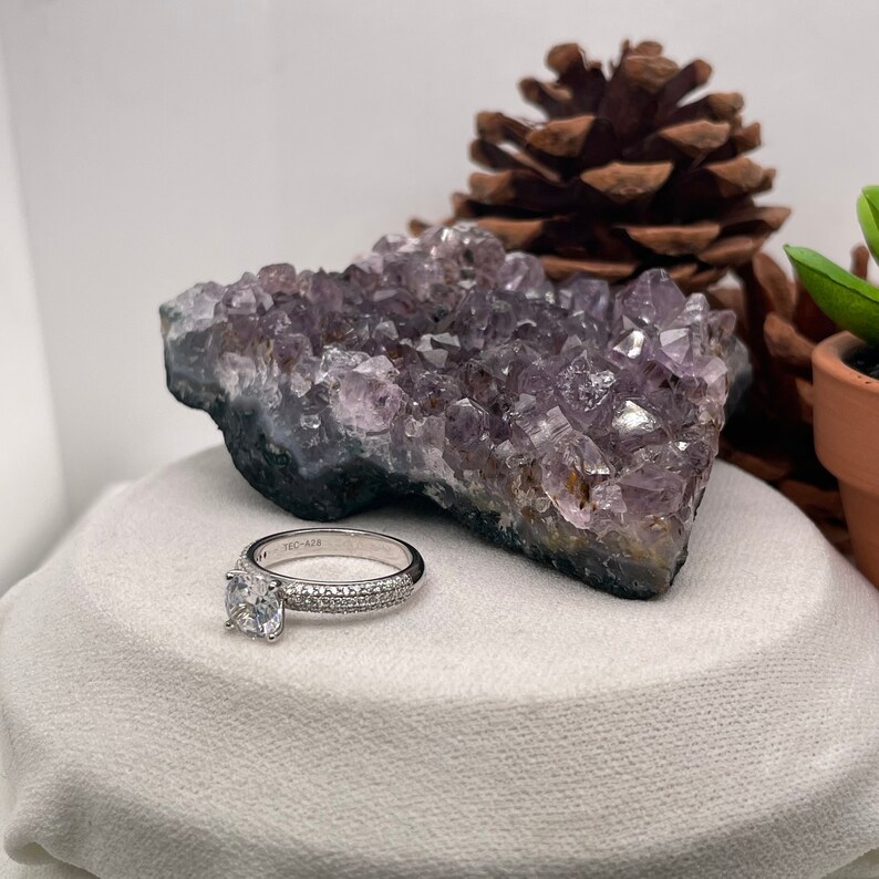 May include: A silver engagement ring with a round centre stone and a band of small diamonds. The ring is sitting on a white cushion with a purple amethyst crystal behind it.