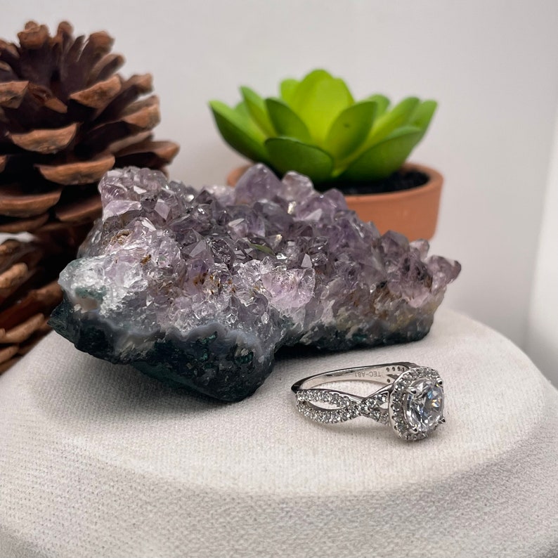 Puede incluir: Un anillo de compromiso de plata con una piedra central redonda y un engaste halo. El anillo tiene una banda retorcida con piedras m&aacute;s peque&ntilde;as engastadas a lo largo de la banda. El anillo se muestra sobre una superficie blanca con un cristal de amatista p&uacute;rpura.