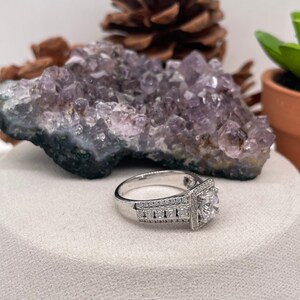 May include: A silver engagement ring with a square halo setting and a large round center stone. The ring is set with smaller diamonds around the band.