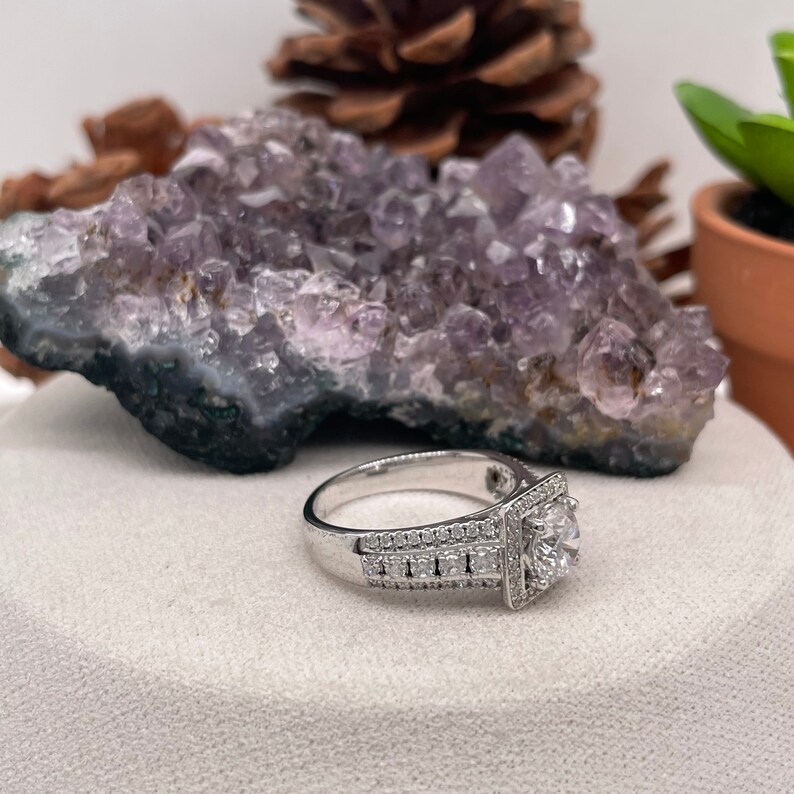 May include: A silver engagement ring with a square halo setting and a large round center stone. The ring is set with smaller diamonds around the band.