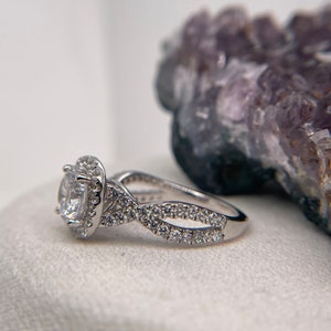 Puede incluir: Un anillo de compromiso de plata con una piedra central redonda rodeada de piedras m&aacute;s peque&ntilde;as. La banda tiene un dise&ntilde;o trenzado con piedras m&aacute;s peque&ntilde;as engastadas a lo largo de la banda.