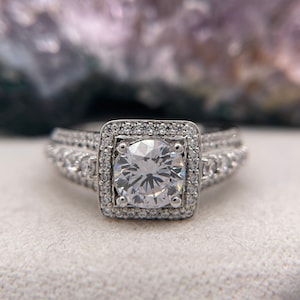 May include: A silver engagement ring with a large, round, clear center stone surrounded by a double halo of smaller stones. The ring has a band with two rows of smaller stones.