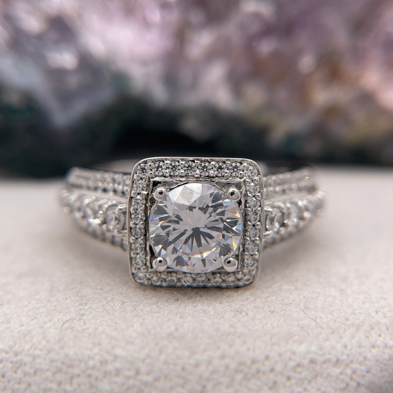 May include: A silver engagement ring with a large, round, clear center stone surrounded by a double halo of smaller stones. The ring has a band with two rows of smaller stones.