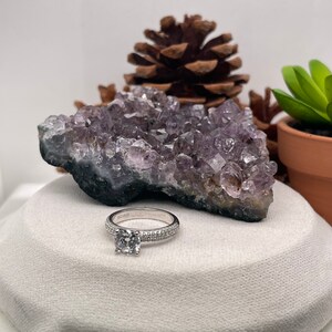May include: A silver ring with a round, clear gemstone surrounded by small, clear gemstones. The ring is on a white surface with a purple amethyst cluster and a pine cone in the background.