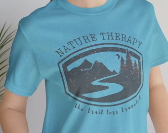 May include: A turquoise t-shirt with a graphic that reads "NATURE THERAPY" above a mountain and river scene. The text "the trail less traveled" is below the graphic. The shirt is made of a soft material.