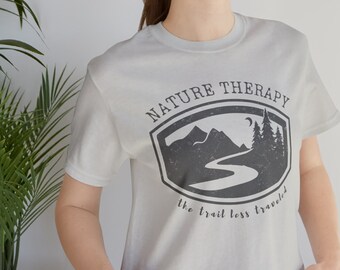 May include: A light gray t-shirt featuring a graphic with the words "NATURE THERAPY" above a mountain and river scene. The text "the trail less traveled" is below the graphic. The shirt is made of a soft material.