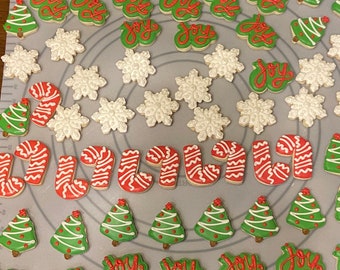 May include: A collection of holiday-themed sugar cookies arranged on a gray silicone mat. The assortment includes red and white candy canes, white snowflakes, green Christmas trees with white icing details, and green cookies with the word joy.