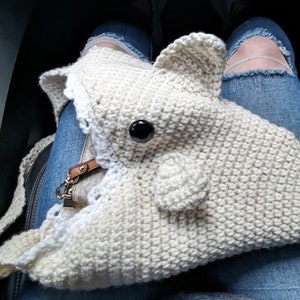 May include: A cream-colored, crocheted shark-shaped bag with a black eye and a small, brown leather strap. The bag is resting on a person's lap, who is wearing blue jeans with ripped knees.