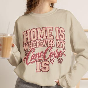 May include: A beige crewneck sweatshirt featuring the red and white text "HOME IS WHEREVER MY Cane Corso IS" with small heart and paw print graphics. The design uses a mix of block and script fonts, creating a clear, centred graphic on the chest.