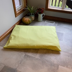 May include: A rectangular pet bed with a light green cover and a white polka dot pattern. The bed is placed on a tiled floor near a window. The bed is designed for a dog or cat.