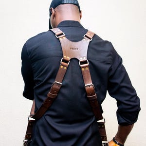 Puede incluir: Un arnés de cuero marrón con hebillas metálicas se lleva sobre una camisa negra. El arnés tiene correas ajustables y una pieza central con la palabra "FEMBY" en relieve. La persona lleva una gorra negra.