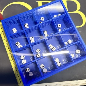 May include: A blue plastic tray with numbered compartments filled with white dice. The tray is open and the dice are visible.