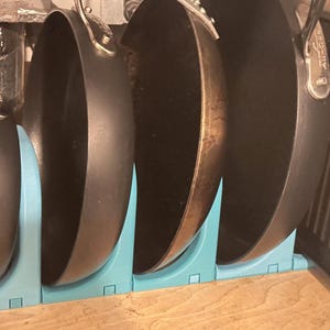 Puede incluir: Cuatro sartenes negras están colgadas de un estante de metal azul. Las sartenes son de diferentes tamaños y tienen un revestimiento antiadherente.