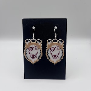 May include: A pair of dog-themed earrings. Each earring features a tan and white dog face with maroon accents and silver-colored ear hooks. The earrings are displayed on a black card against a white background.