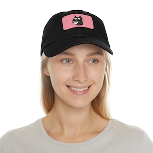 Peut inclure: Une casquette de baseball noire avec un écusson rectangulaire rose sur le devant. L'écusson présente une illustration graphique d'un chat noir aux yeux écarquillés tenant une cuillère blanche et mangeant dans un petit bol blanc sur fond rose.