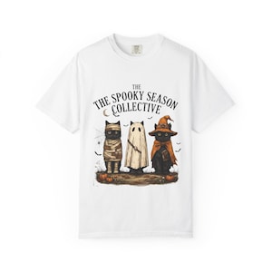May include: A white short-sleeve t-shirt featuring a graphic of three black cats in Halloween costumes, including a mummy, a ghost, and a witch. The text above reads THE SPOOKY SEASON COLLECTIVE. The design includes small pumpkins and bats.