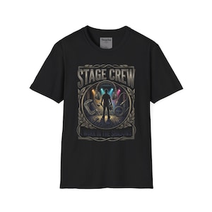 May include: A black short-sleeved t-shirt featuring a graphic print with the text STAGE CREW and I WORK IN THE SHADOWS. The design depicts a silhouette of a person surrounded by stage equipment, including headphones, cables, and spotlights in blue and orange.