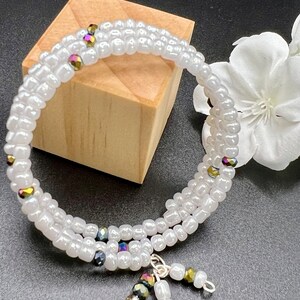 May include: A triple-wrap bracelet made with white beads and iridescent accents. The bracelet has a silver charm with three dangling iridescent beads.