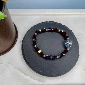 May include: A red crystal bracelet with a clear crystal heart charm. The bracelet is on a black slate coaster.