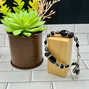 Puede incluir: Pulsera de cuentas negras y blancas con un dije de borla negro. La pulsera está en un expositor de madera.