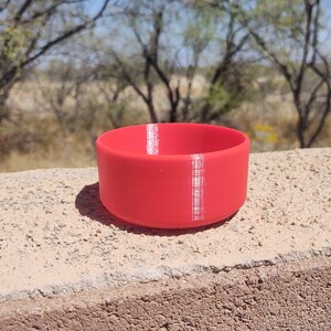 Pu&ograve; includere: Una piccola ciotola cilindrica rossa con una striscia bianca lungo il lato. La ciotola &egrave; realizzata in un materiale liscio, simile alla plastica.