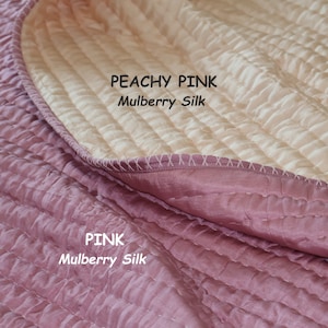 Può includere: Primo piano di un piumino in seta di gelso rosa pesca e rosa. Il piumino è trapuntato con un motivo a rombi e ha una lucentezza sottile. Il testo "PEACHY PINK Mulberry Silk" e "PINK Mulberry Silk" è visibile sul piumino.