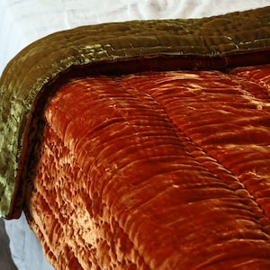 Puede incluir: Primer plano de un edredón acolchado. El edredón presenta un color naranja quemado cálido en el cuerpo principal, contrastado por un borde verde oliva oscuro. La tela tiene un brillo suave y aterciopelado, lo que sugiere una sensación lujosa. El acolchado crea una superficie texturizada.