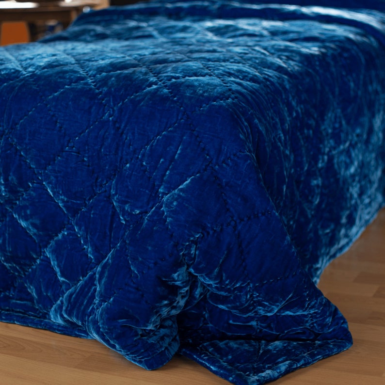 May include: A blue velvet quilted throw blanket with a diamond pattern. The blanket is folded and draped over a wooden surface.
