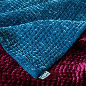 May include: A teal blue velvet quilted throw blanket folded over a burgundy velvet quilted throw blanket. The teal blue blanket has a tag that reads "Luna's Choice".
