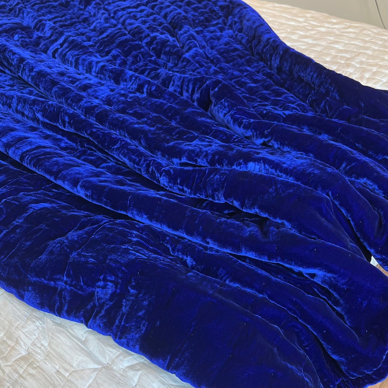 K&ouml;nnte beinhalten: Eine blaue, gesteppte Decke aus Samt mit weicher, pl&uuml;schiger Textur. Die Decke ist gefaltet und &uuml;ber eine wei&szlig;e Oberfl&auml;che gelegt.