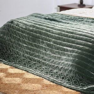 May include: A green velvet quilted throw blanket with a subtle sheen. The blanket is folded and draped over a bed with a woven rug underneath.