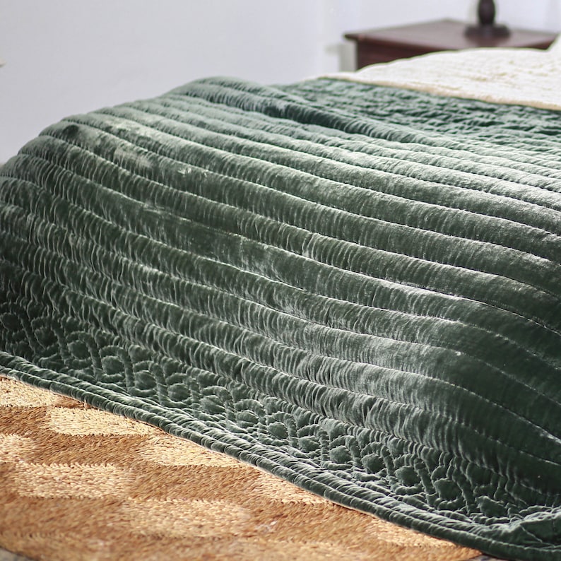 May include: A green velvet quilted throw blanket with a subtle sheen. The blanket is folded and draped over a bed with a woven rug underneath.