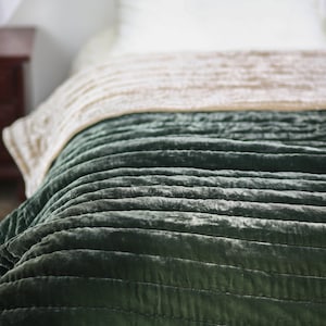 May include: A green velvet quilted throw blanket with a subtle sheen. The blanket is folded and draped over a bed.