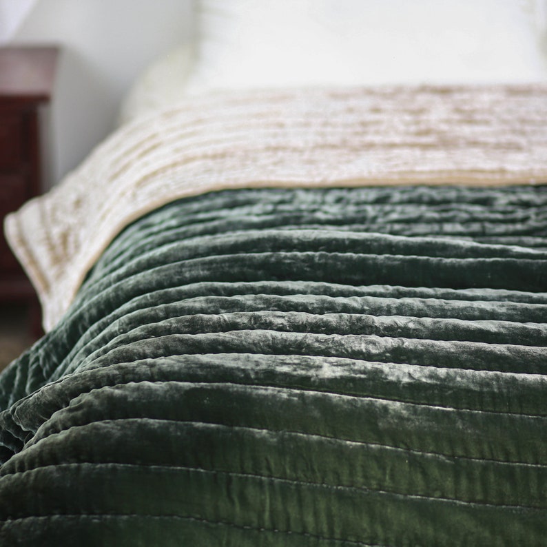 May include: A green velvet quilted throw blanket with a subtle sheen. The blanket is folded and draped over a bed.