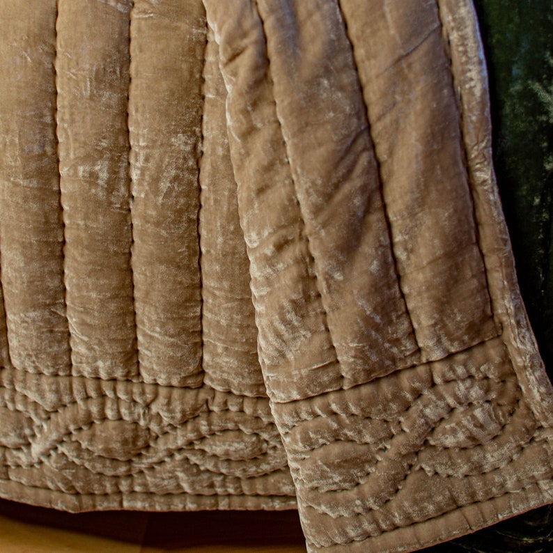 May include: A close-up of a beige quilted throw blanket with a textured, velvety surface. The blanket is folded and has a decorative stitch pattern around the edges.