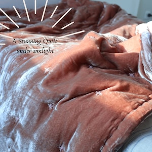 May include: A close-up of a soft, blush-coloured velvet quilt with a subtle star pattern. The quilt is folded and draped over a bed, with sunlight illuminating the fabric.