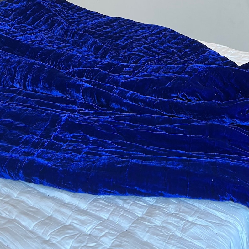 K&ouml;nnte beinhalten: Eine k&ouml;nigsblaue Samtdecke mit Steppmuster. Die Decke ist gefaltet und &uuml;ber ein wei&szlig;es Bett gelegt.