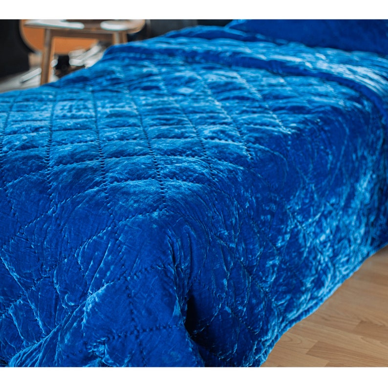 May include: A blue velvet quilted throw blanket with a diamond pattern. The blanket is folded and draped over a bed.