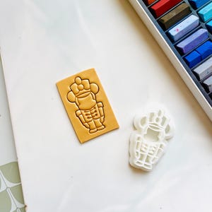 May include: A white plastic cookie cutter shaped like a nutcracker, next to a brown clay impression of the same design.