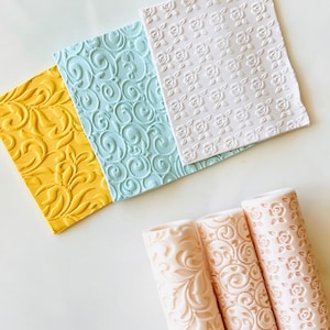 May include: Three rolling pins with different embossed patterns, including floral and swirl designs. The rolling pins are beige and made of plastic. Three square textured sheets with different patterns are also shown in the image. The sheets are yellow, blue, and white.