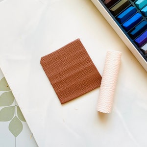 May include: A brown square of clay and a white rolling pin on a white surface. The clay has a textured surface.