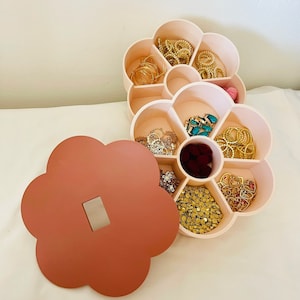 Könnte beinhalten: Ein blütenförmiger Schmuck-Organizer mit hellrosa Basis und passendem Deckel. Der Organizer ist mit verschiedenen goldfarbenen Schmuckstücken gefüllt, darunter Halsketten, Armbänder und Ohrringe. Der Deckel hat eine gedämpfte rotbraune Farbe.