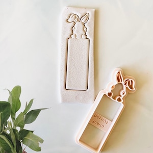 May include: A pink plastic cookie cutter in the shape of a bunny rabbit holding a blank bookmark. The bookmark has the text "pernaiz Jewel" on it.