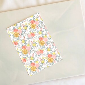 May include: A white background with a repeating pattern of pink, yellow, and gray flowers. The flowers are arranged in a random pattern and are surrounded by green leaves.